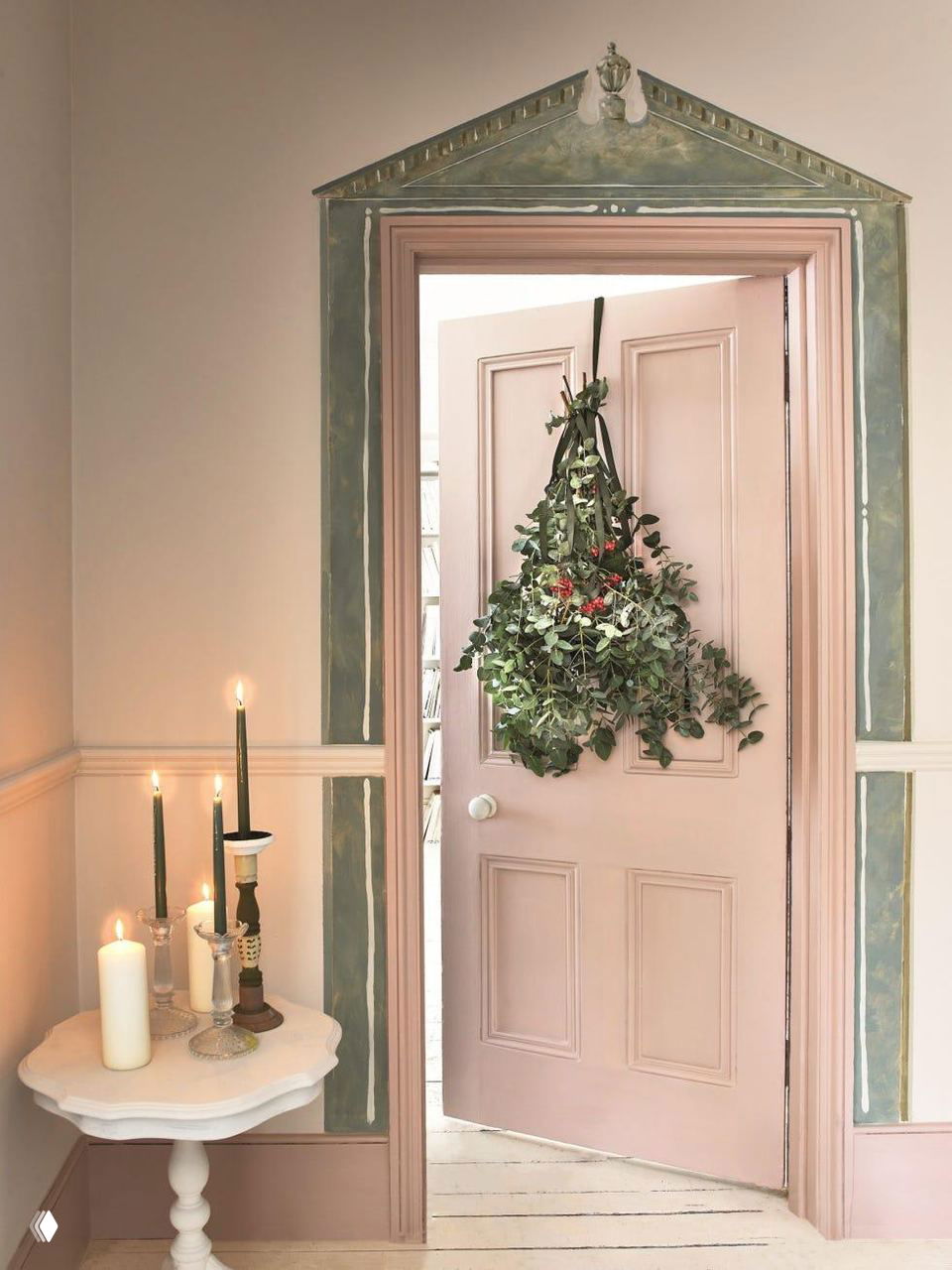 Нежно-розовая дверь, украшенная подвесной композицией из эвкалипта, хвои и ягод; рядом столик со свечами создаёт мягкий праздничный свет.
