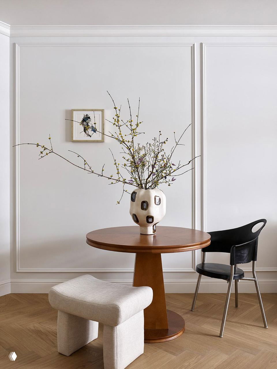 Минималистичный угол столовой: круглый деревянный стол, необычная керамическая ваза с ветвями и сочетание современных стульев и мягкого пуфа на фоне лепнины.