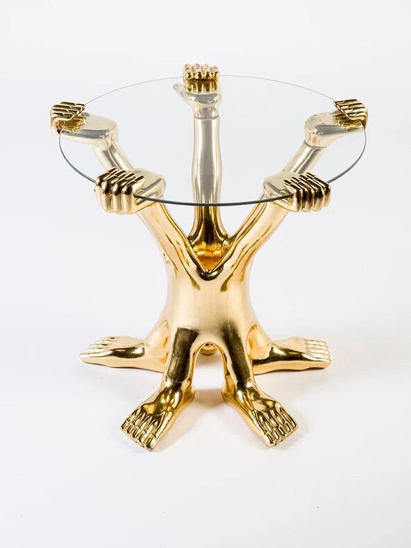 Низкий стол со стеклянной столешницей, опирающейся на золотые фигурные опоры в виде человеческих форм, дизайн с сюрреалистическим акцентом на деталях.