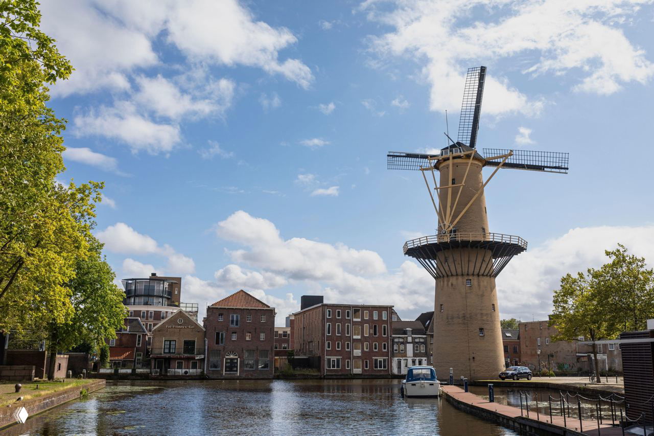 Ветряная мельница у канала в европейском городе, её отражение в воде, прогулочные причалы и низкая застройка под облачным небом.