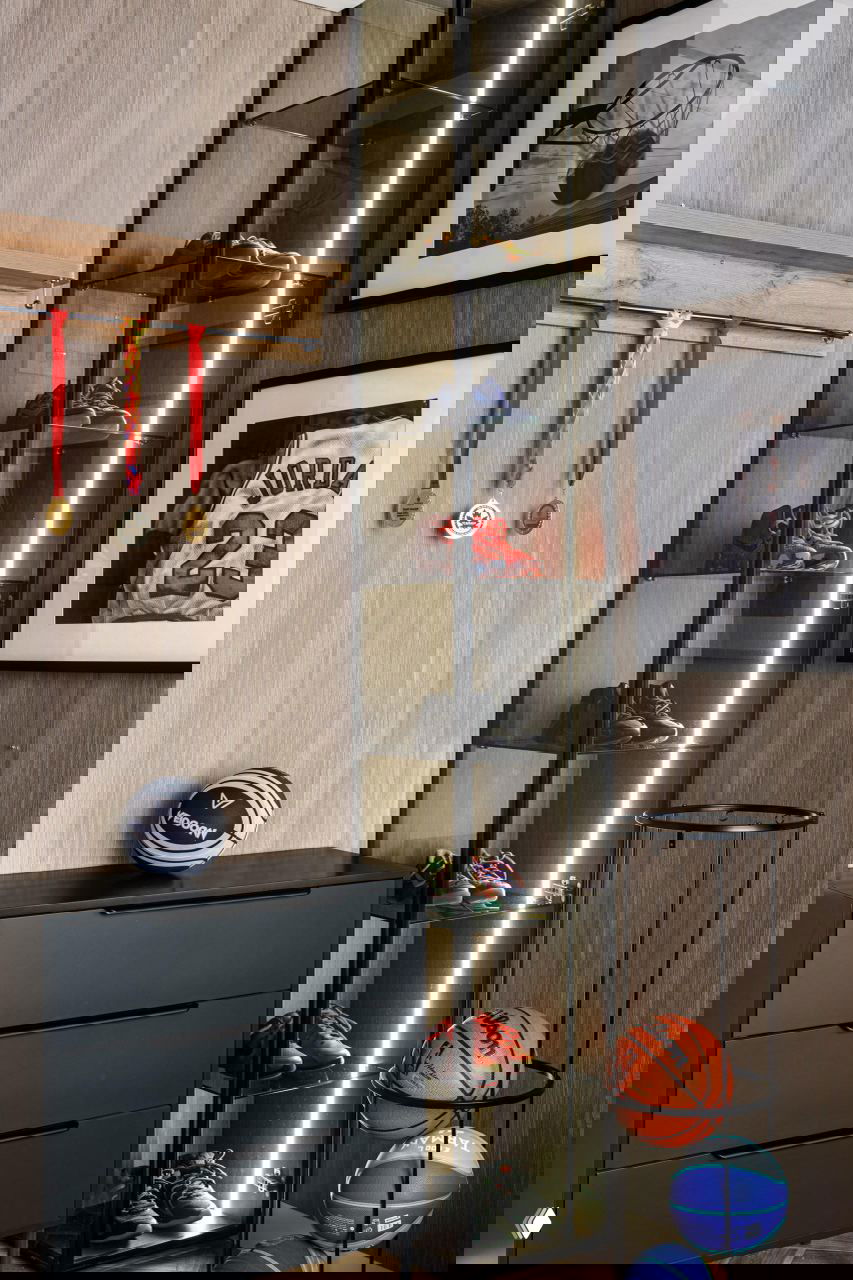 Спортивный уголок в квартире: витрина со стеклянными полками и кроссовками, настенные медали и баскетбольные мячи, чёрный комод и постеры с баскетбольной тематикой.