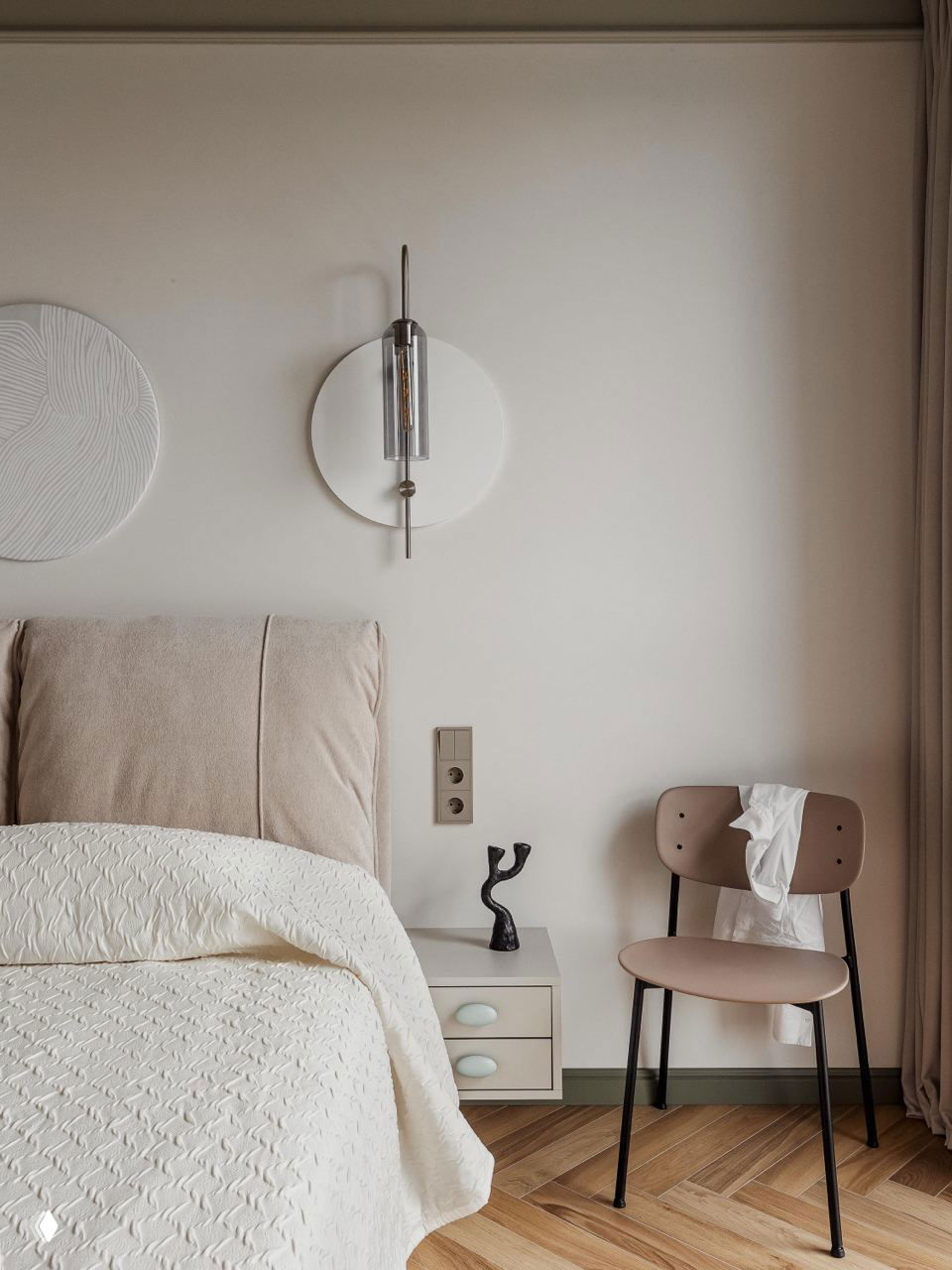 Уютный уголок спальни: мягкое изголовье, светлая постель, настенный светильник в металлическом корпусе и простая деревянная тумбочка на фоне нейтральной стены и паркета.