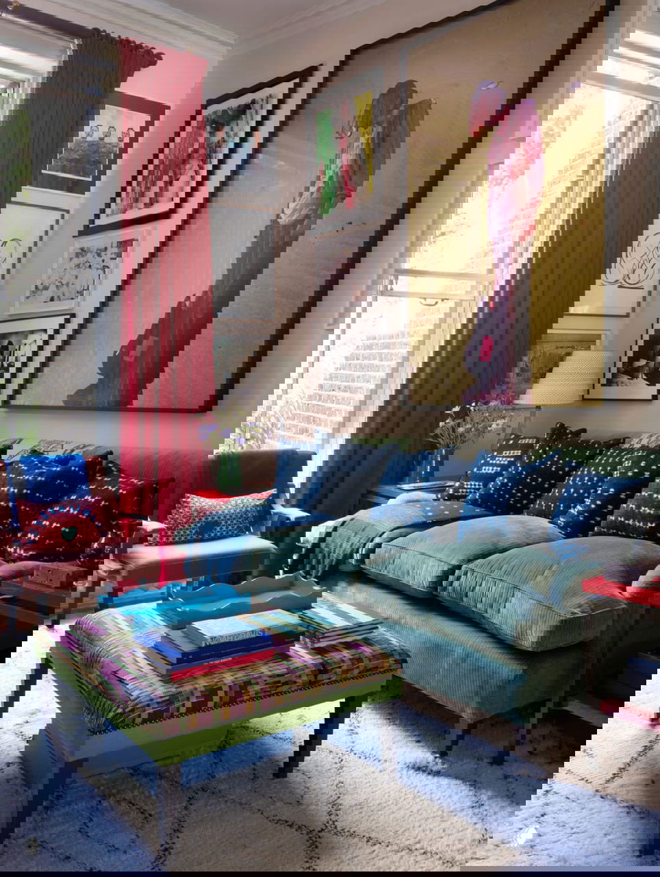 Гостиная с зелёным диваном, высокой красной занавесью, галереей рамок на стене и разноцветным пуфом на переднем плане — слои текстур и картин.