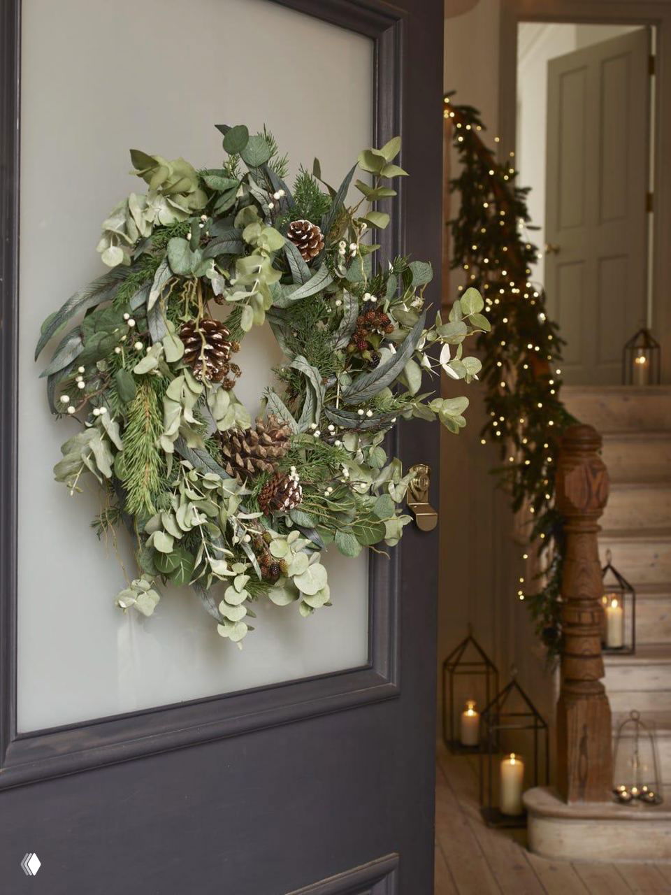 Дверной венок из хвои, эвкалипта и крупных шишек на тёмной дверной панели; в глубине кадра заметна подсвеченная гирлянда у лестницы и свечи.