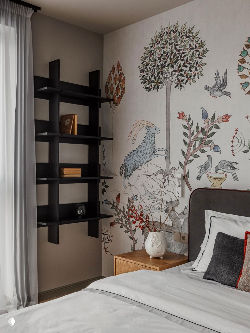 Деталь спальни с настенной росписью: растительные и животные мотивы, полка‑стеллаж чёрного цвета и прикроватная тумбочка с керамической вазой и декором.