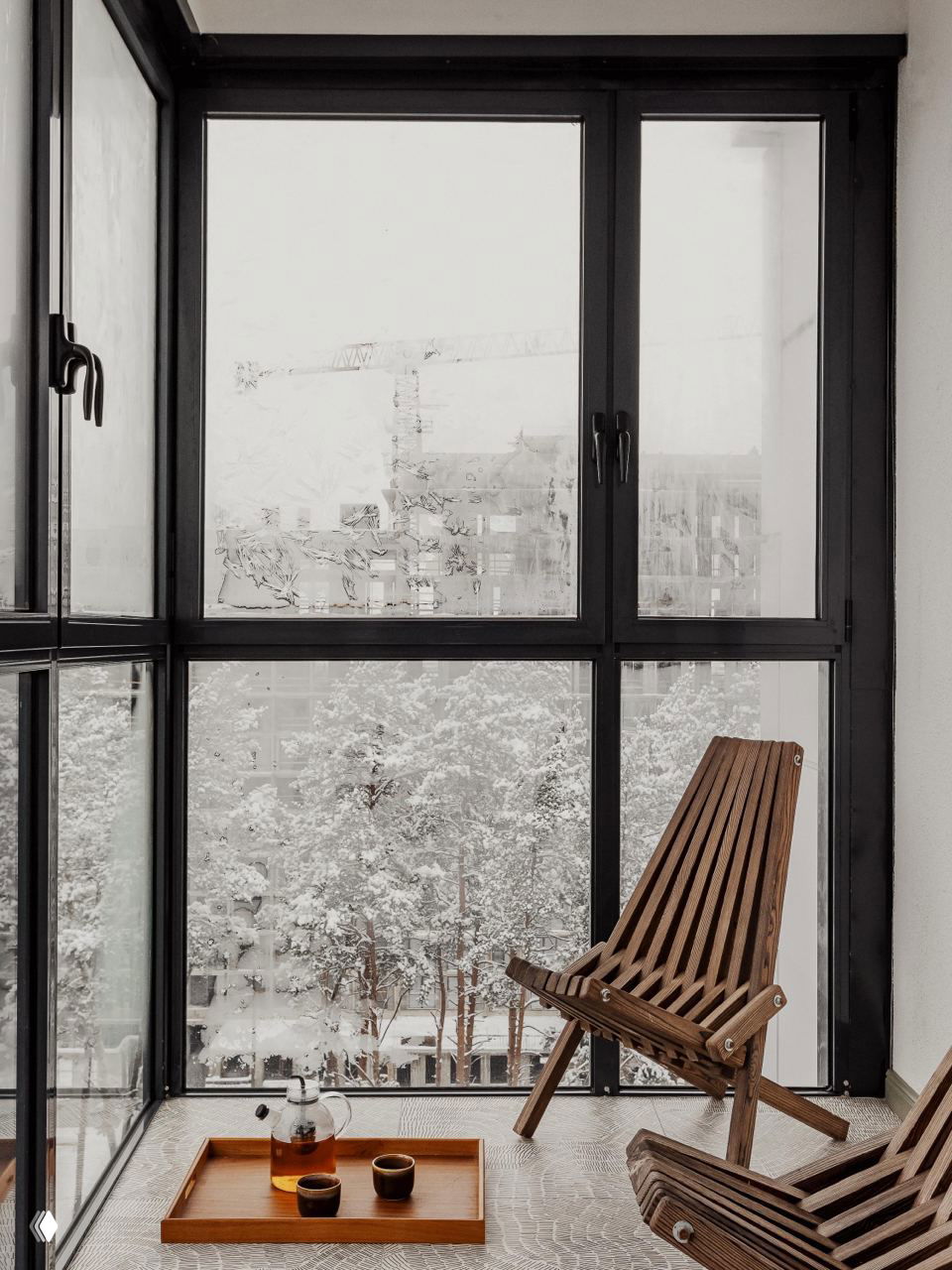 Лоджия с панорамными тёмными рамами и видом на заснеженный лес; деревянные раскладные кресла и поднос с чайником создают атмосферу уединения и уюта.