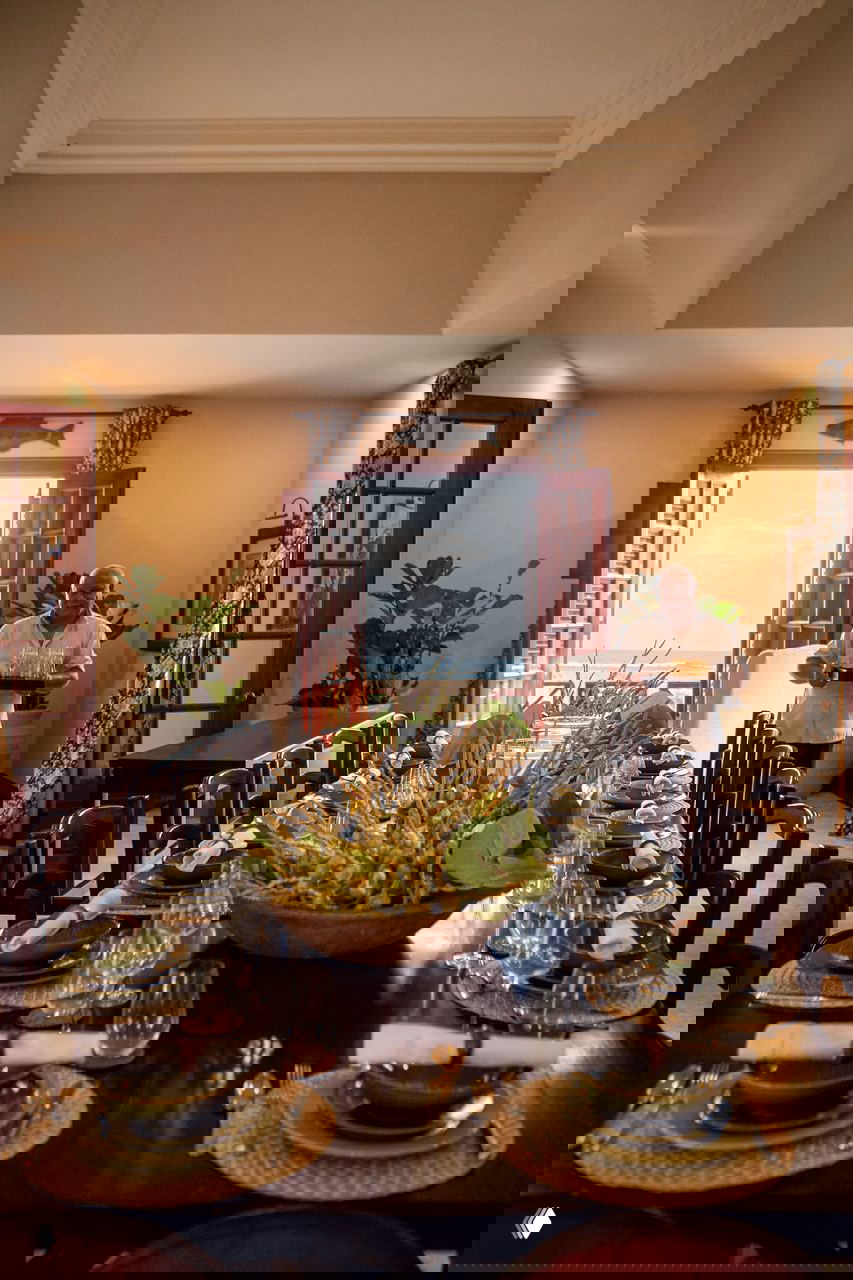 Обеденная зона в доме в Доминикане: длинный стол, сервировка, центральная растительная композиция и вид на океан через открытые ставни.