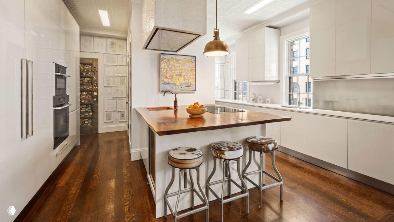 Кухня с островом и барными стульями, белая кухня в сочетании с деревянной столешницей острова, окна с видом на город и аккуратная планировка рабочей зоны.