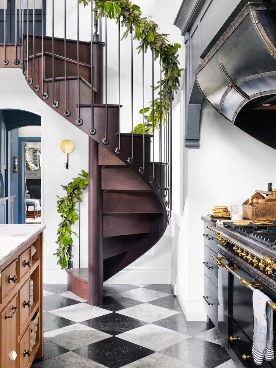 Кухонный интерьер с винтовой деревянной лестницей: перила украшены свежей зеленью, контрастирующей с чёрно‑белой плиткой и интерьерными деталями.
