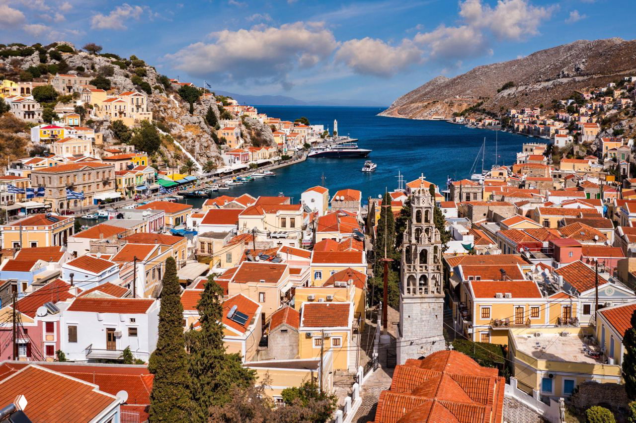 Панорамный вид на прибрежный город с красочными домами у гавани, яхтами и узким морским проливом в окружении скал и террас.