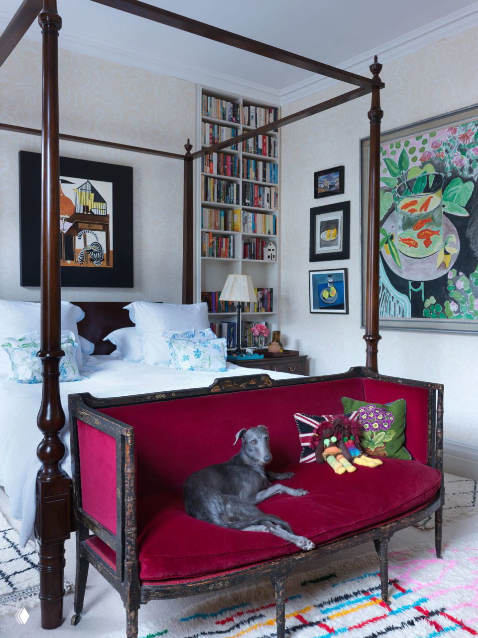 Спальня с четырёхстоечной кроватью, картинами на стенах и бархатным красным диваном в изножье, на котором лежит собака — уют и семейная атмосфера.