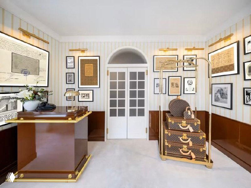 Комната‑«номер» в стиле архивного кабинета Louis Vuitton: стопка монограммированных чемоданов на тележке, винтажные плакаты и оформленные стеновые панели.