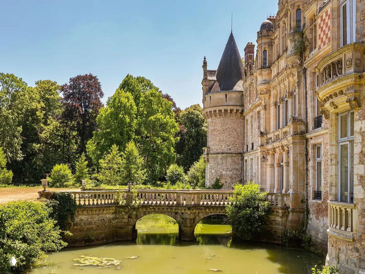 Боковой фасад замка Эсклимон с каменным мостом через ровой пруд, детализированная отделка и зелёная растительность вокруг.