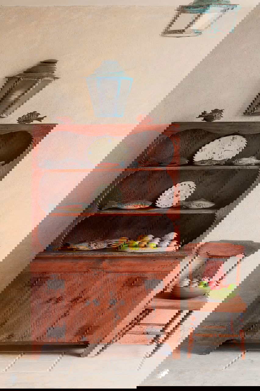 Антикварный деревянный шкаф с коллекцией посуды и декоративными предметами в доме в Доминикане; рядом простая деревянная мебель и корзина.