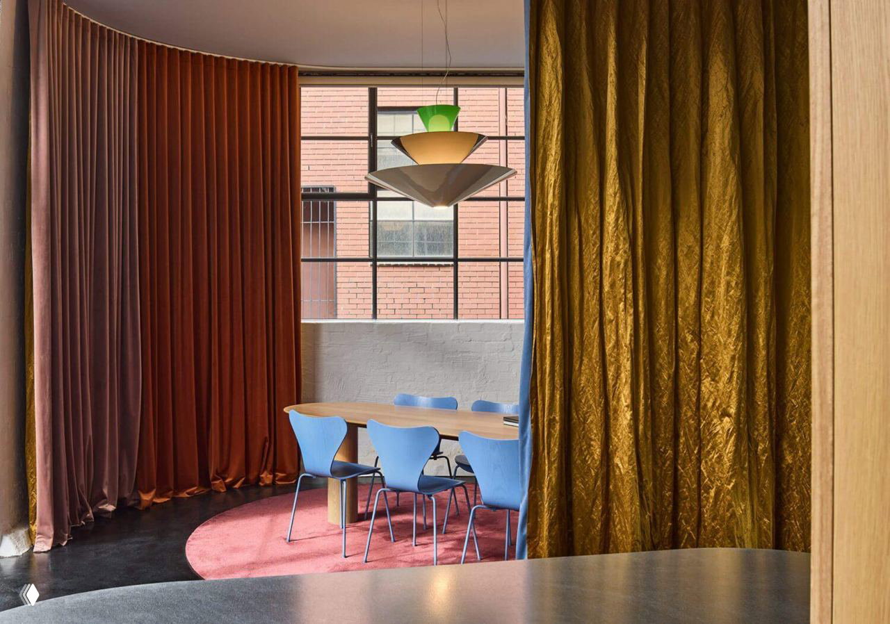 Вид через бархатные занавеси на столовую/переговорную студии: крупный подвесной светильник, высокое промышленное окно, кирпичная стена и игра текстур тканей.