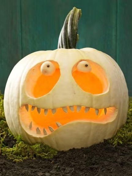 Бледная белая тыква с круглыми глазами и зубастым ртом, подсвеченная изнутри; изображение показывает фактуру поверхности и контраст между светом и темной почвой рядом.