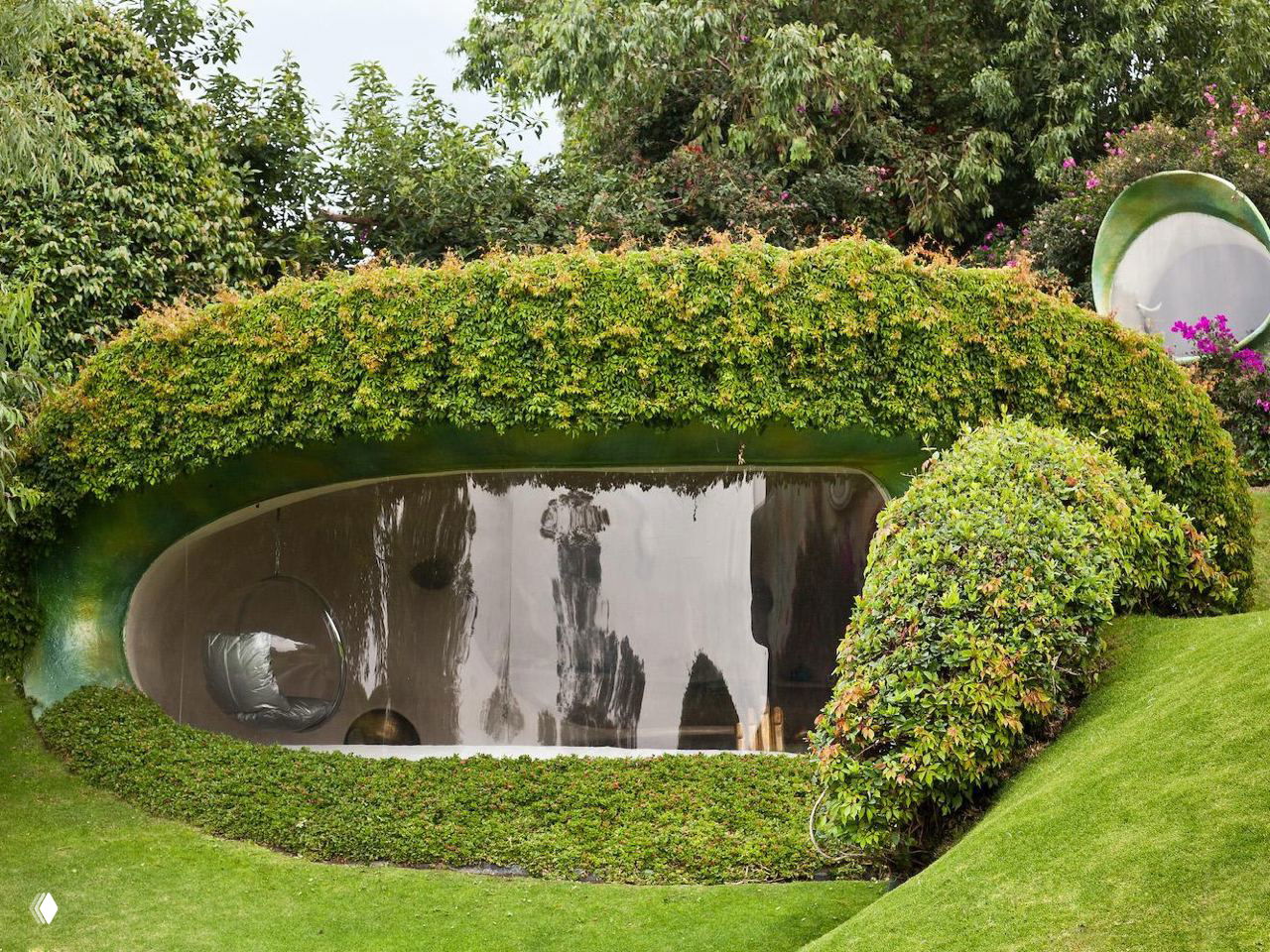 Фрагмент фасада дома врезанного в склон: крупное овальное окно с отражающим стеклом и растительность по краям ниши.