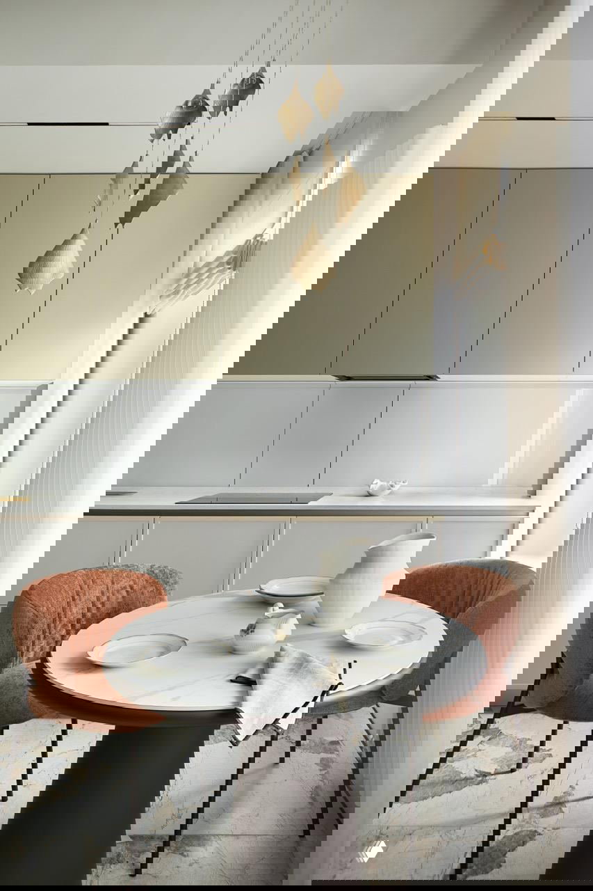 Обеденная зона с круглым столом и бархатными креслами тёплого терракотового оттенка на фоне приглушённой неоклассической кухни и подвесных светильников.