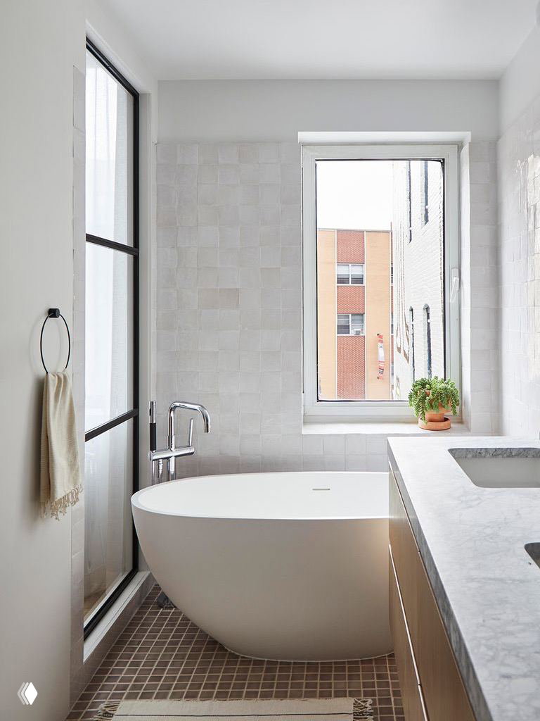 Современная белая ванная с овальной свободностоящей ванной у окна; светлая плитка, подоконник с комнатным растением и мягкая нейтральная палитра.