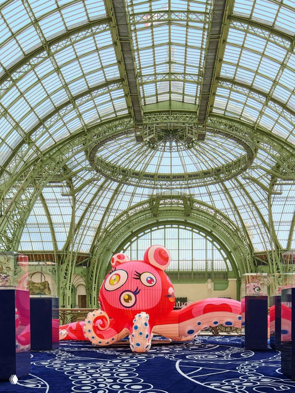 Крупный ракурс на тело и щупальца розово‑красного осьминога, расположенного на синем ковре с узором, видно пространство выставки и архитектуру зала.