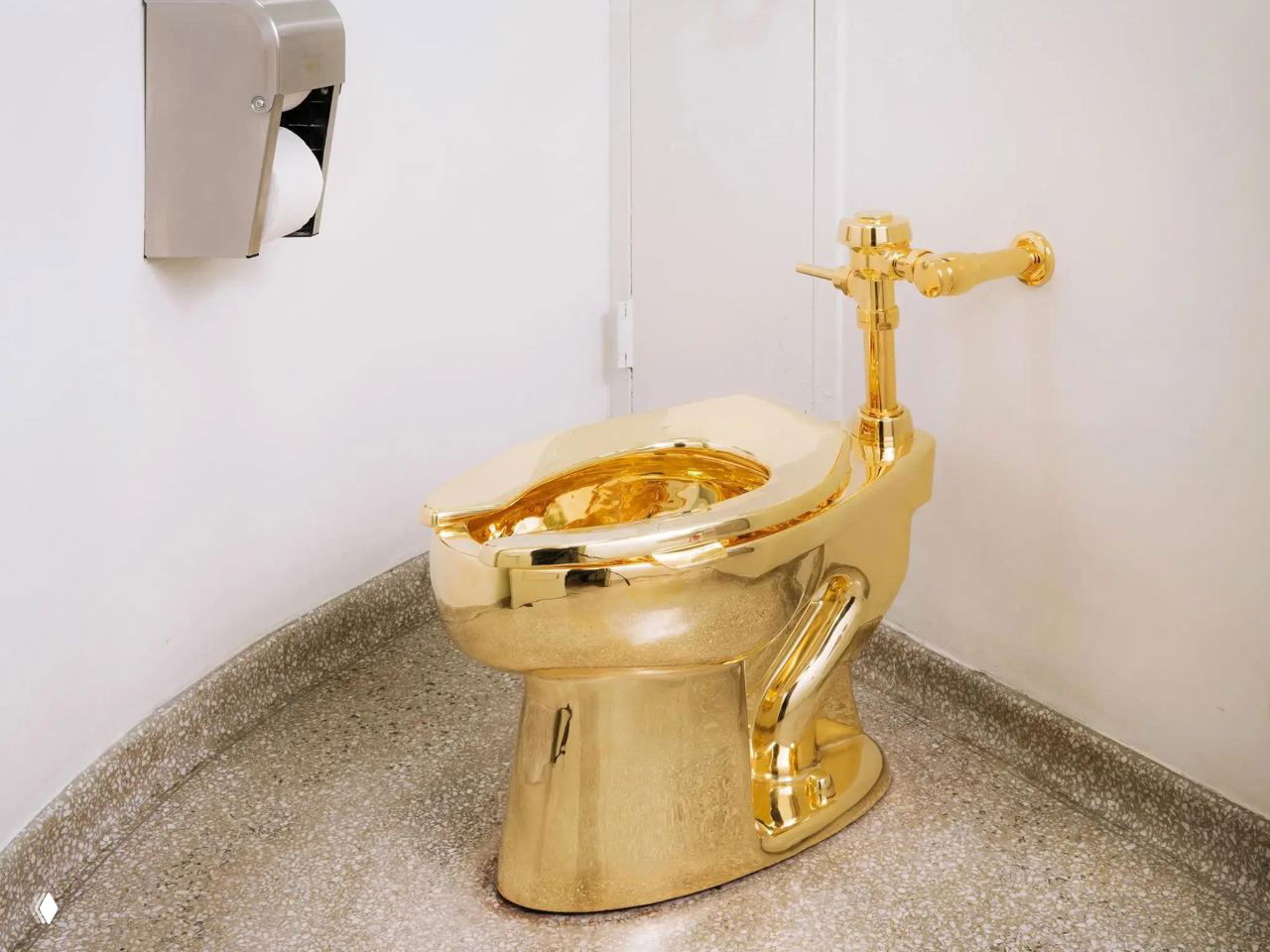 Крупный план блестящего золотого унитаза художника — видно сияющую 18‑каратную поверхность, сливной механизм и настенное держало для бумаги на светлом фоне.