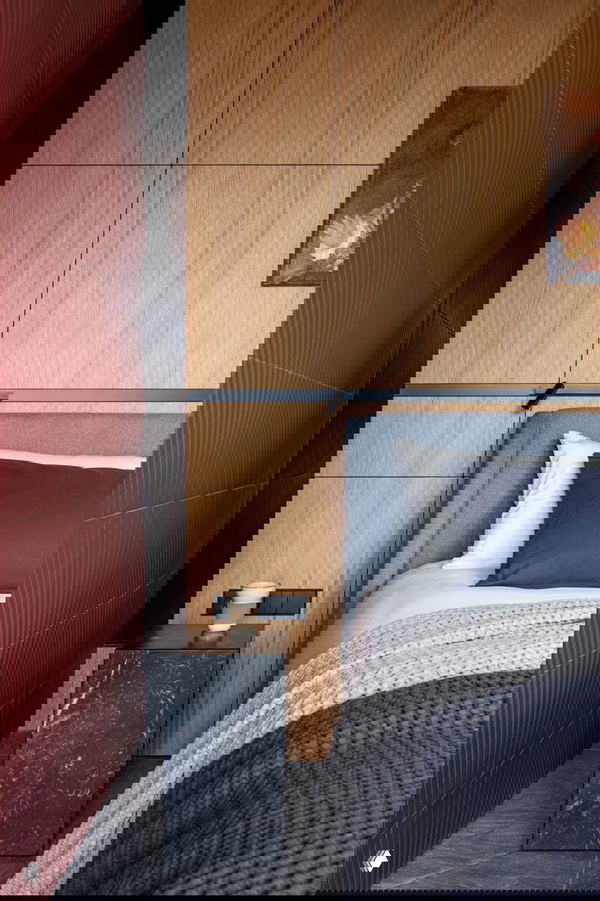 Деталь спальни: деревянные панели за изголовьем, текстурная бордовая стена, прикроватная тумба из черного камня и акцентный текстиль на кровати.