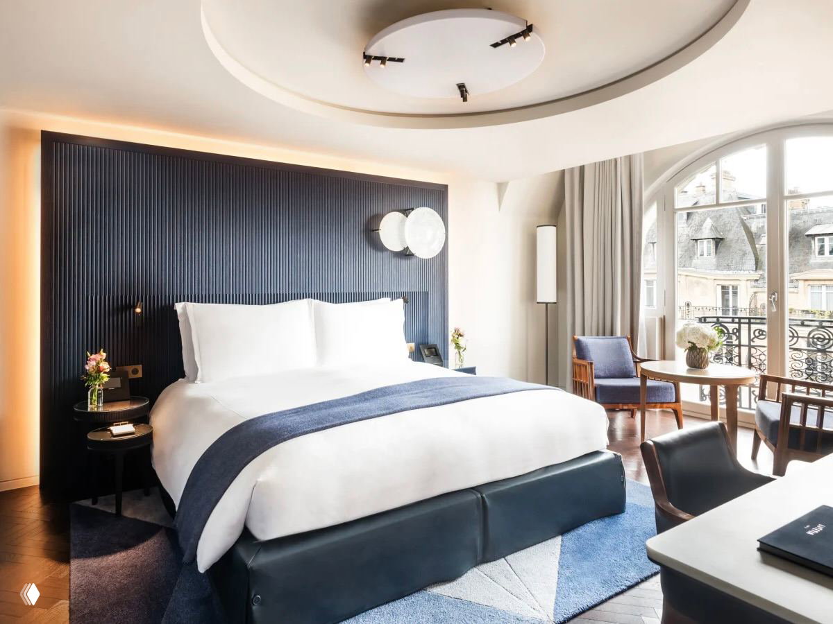 Интерьер номерa Mandarin Oriental Lutetia: кровать с белым постельным бельём, сдержанный современный декор и арочные окна с видом на город.