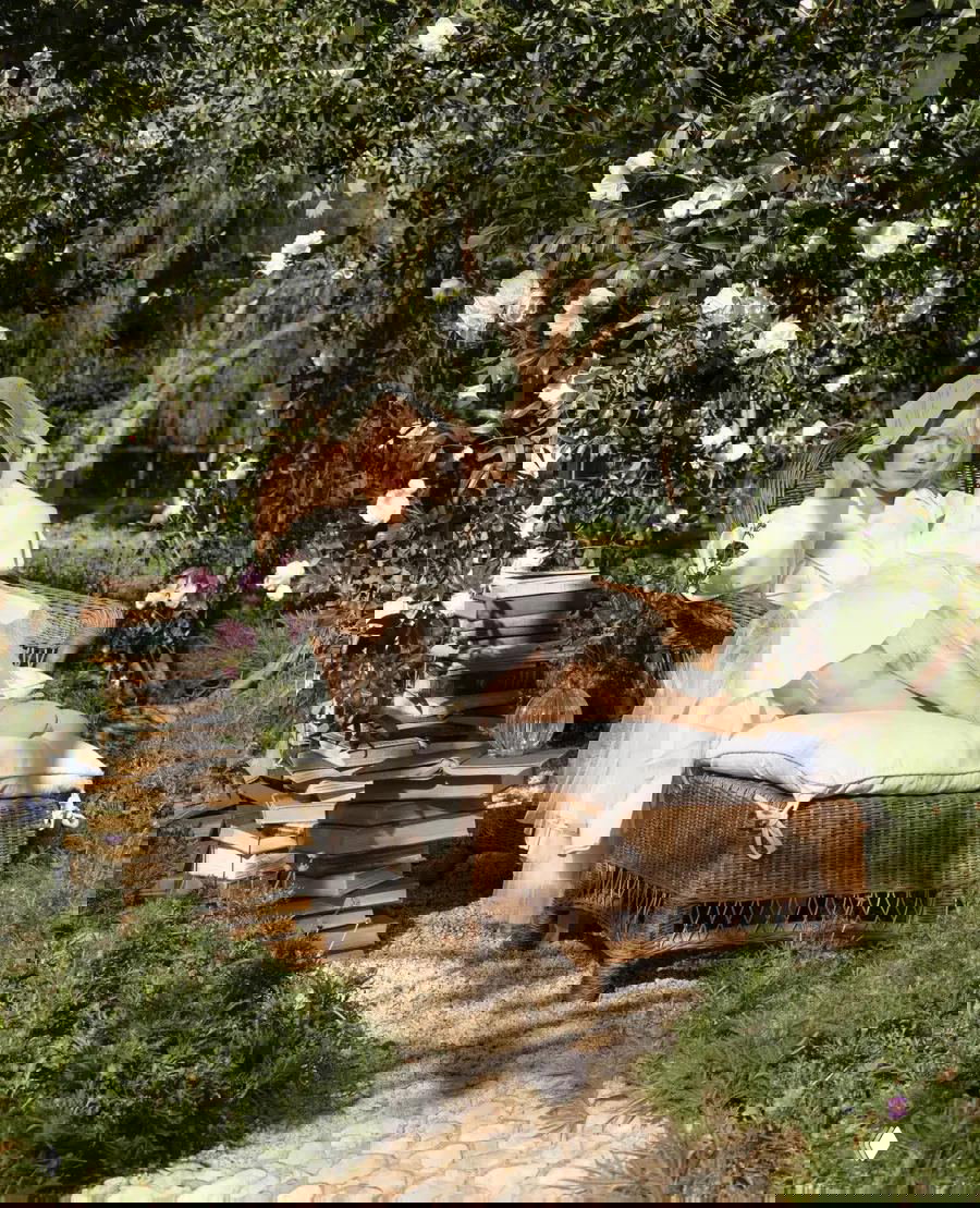 Женщина отдыхает в ротанговом кресле на открытом воздухе под аркой белых цветов, рядом стопка книг и естественный садовый ландшафт.