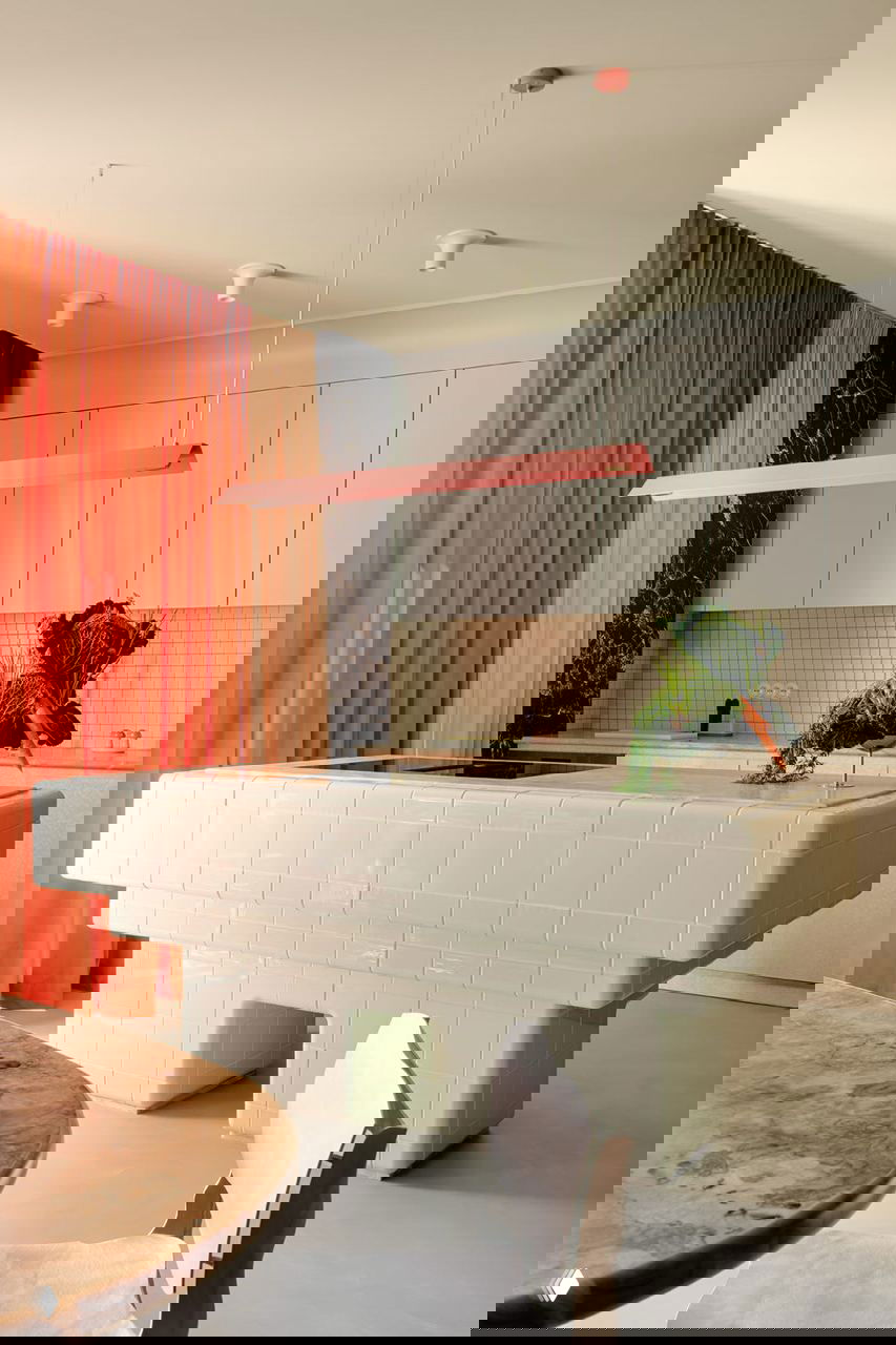 Кухонный остров в облицовке белой плитки скульптурной формы, навесная розовая лампа и минималистичный фон кухонных шкафов.