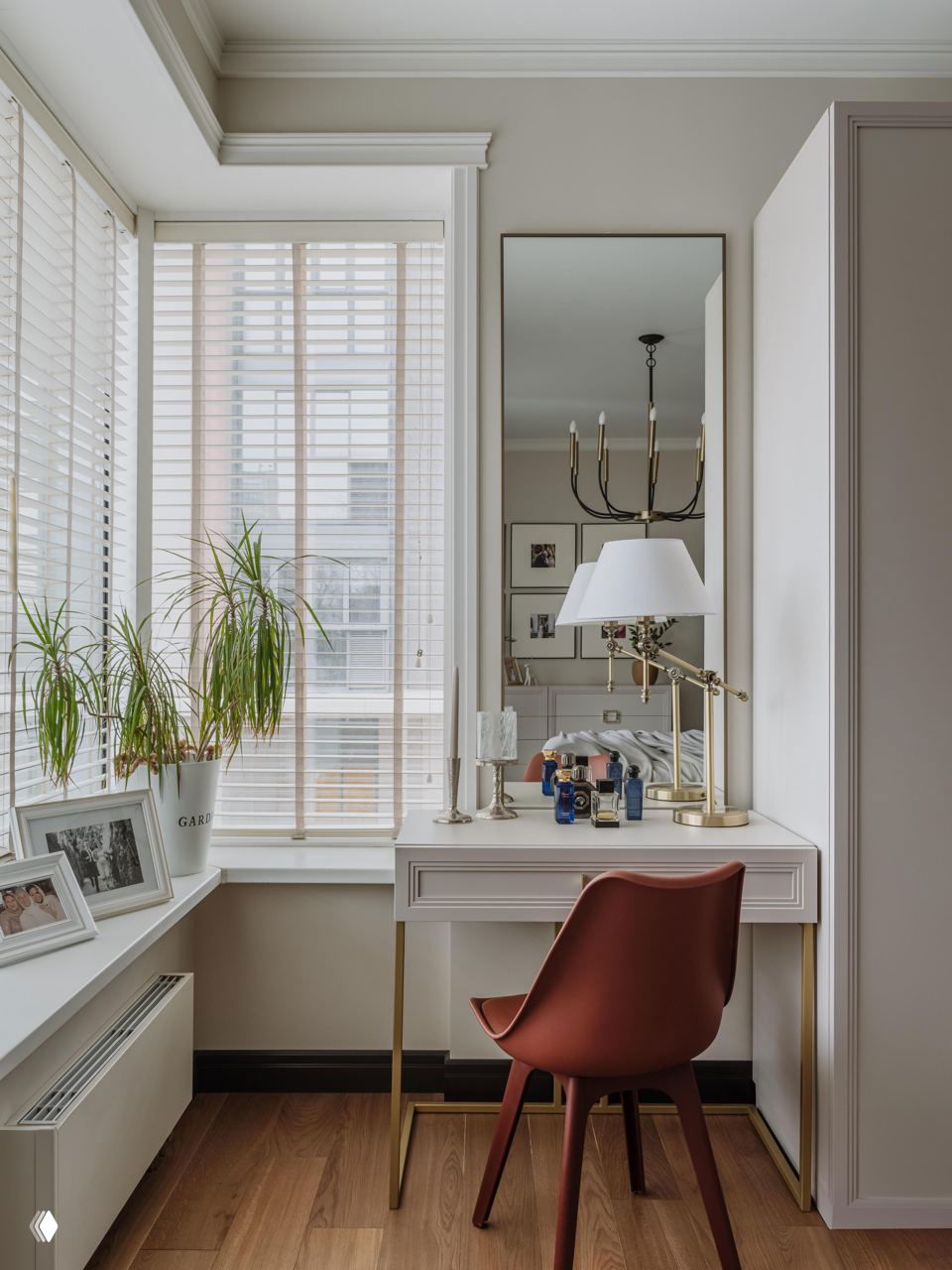 Рабочий уголок у окна: небольшой столик, зеркало в полный рост, кресло терракотового оттенка и белые жалюзи, создающие мягкое дневное освещение.