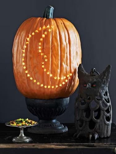 Высокая тыква с рисунком из множества отверстий в виде полумесяца, стоит на подставке рядом с декоративными элементами; контраст отверстий и подсветки создаёт узор.