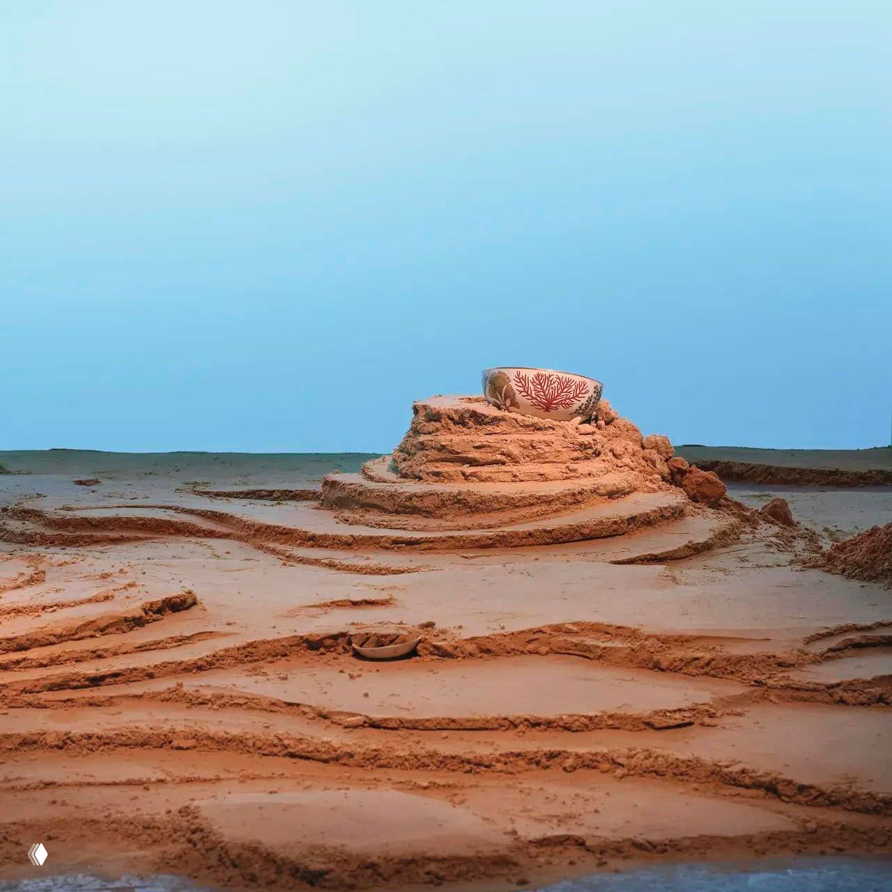 Одиночная чашка Hermès с рисунком морской водоросли, поставленная на песчаную насыпь и снятая на фоне голубого неба инсталляции