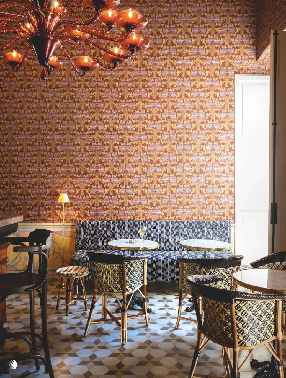 Кафе-зона бутик-отеля: небольшие круглые столики, ротанговые стулья и яркие обои с орнаментом, пол выложен графичной керамической плиткой — атмосферный уголок для гостей.