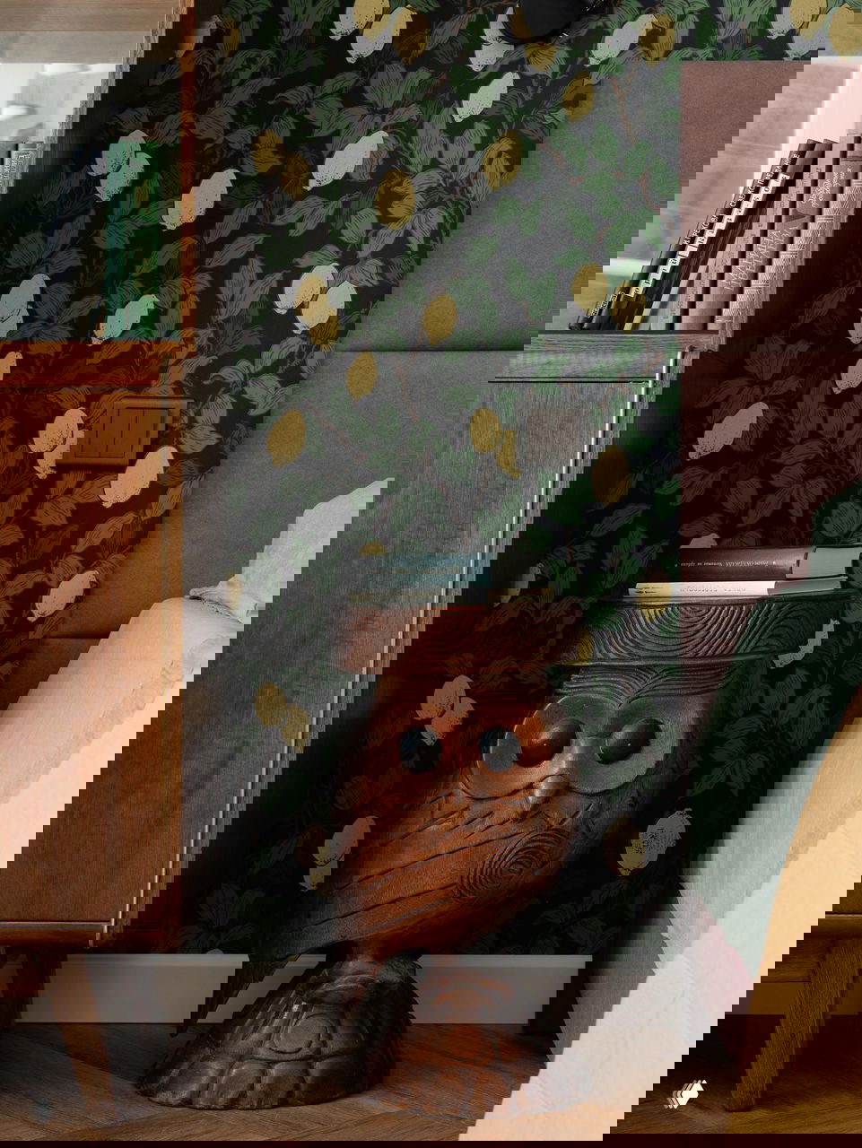 Крупный план резного деревянного табурета-совы рядом с кроватью, на фоне темно-зеленых обоев и открытой витрины с книгами и аксессуарами.