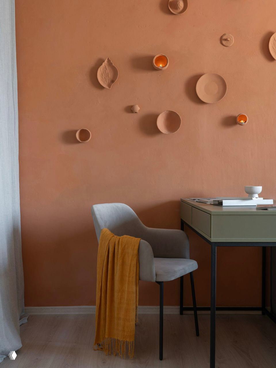 Рабочий уголок у стены, на фоне глубокого терракотового покрытия: серый стул, жёлтый плед и декоративные настенные элементы создают теплую атмосферу