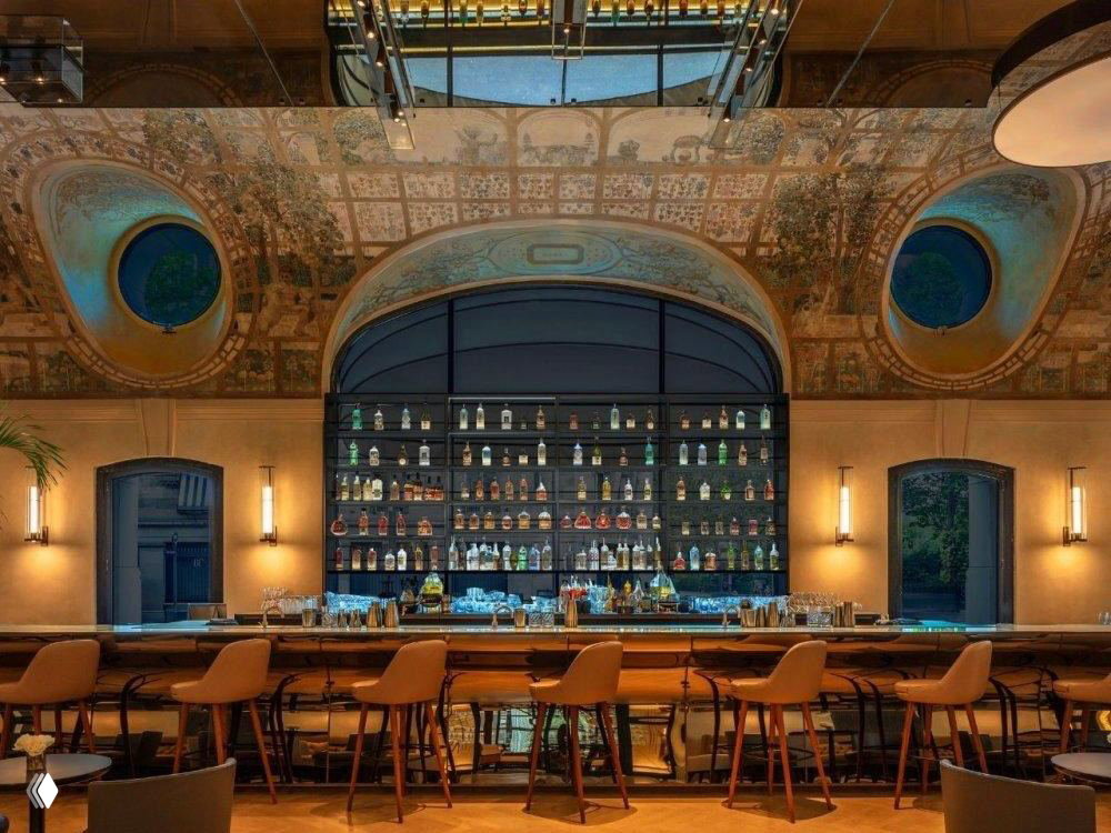 Интерьер бара отеля Mandarin Oriental Lutetia с высокими стульями у длинной стойки, декоративным потолком и тёплой атмосферой вечера.