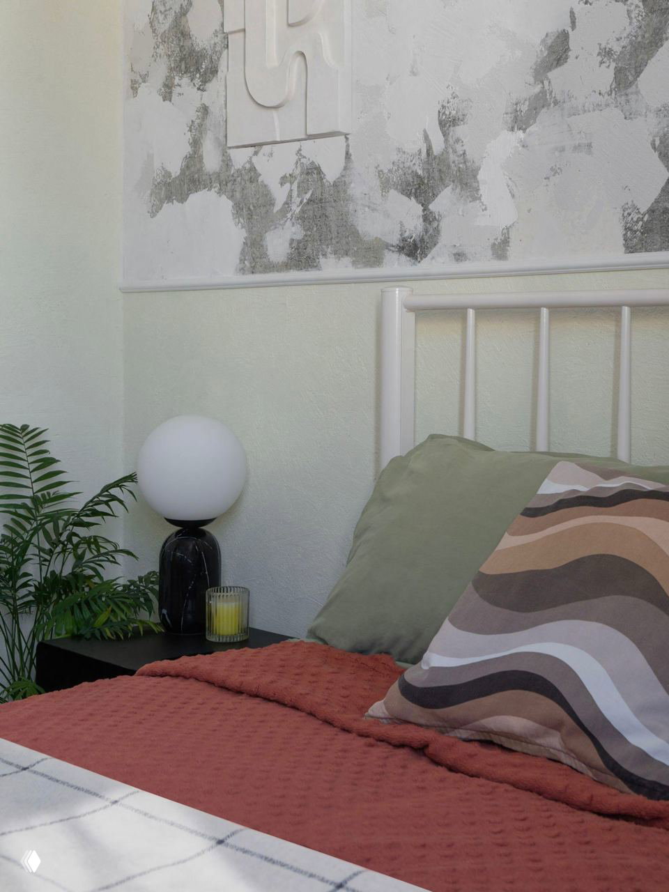 Кровать с текстурным терракотовым пледом и декоративной волнистой подушкой, рядом тумбочка с круглой лампой и растением — мягкие тона и фактуры спальни