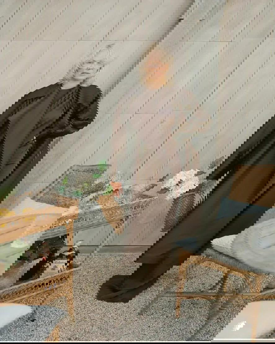 Женщина в тёмном наряде стоит рядом с ротанговой мебелью на террасе, в руках плетёная корзина с цветами, интерьеры в естественной цветовой гамме.