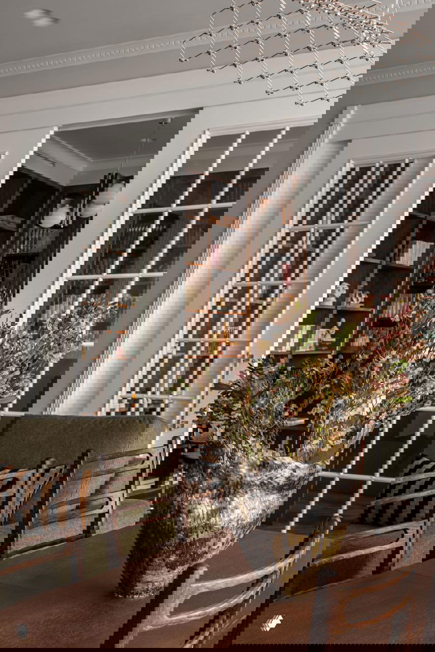 Интерьер со стеклянными распашными дверями, вид на кабинет с книжными полками, на переднем плане стол с декоративной керамической вазой и стульями.