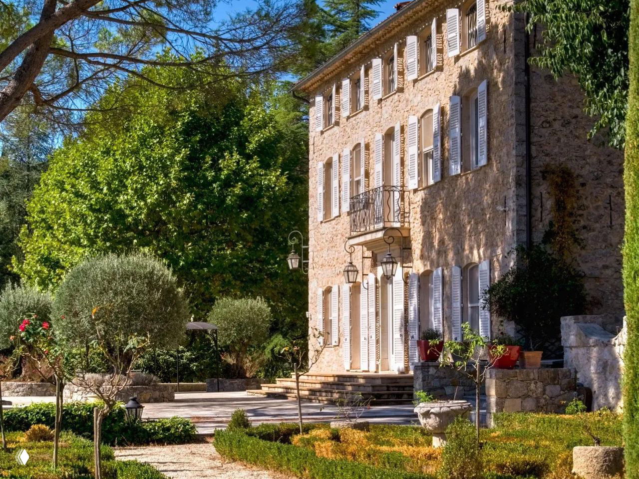 Каменное жилое здание усадьбы с белыми ставнями и маленьким балконом, окружённое ухоженным садом и патио из камня.