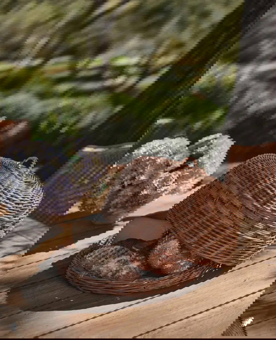 Деревянный стол на открытом воздухе с буханками хлеба под плетёным куполом и вазой с гортензиями, тёплое естественное освещение летнего дня.