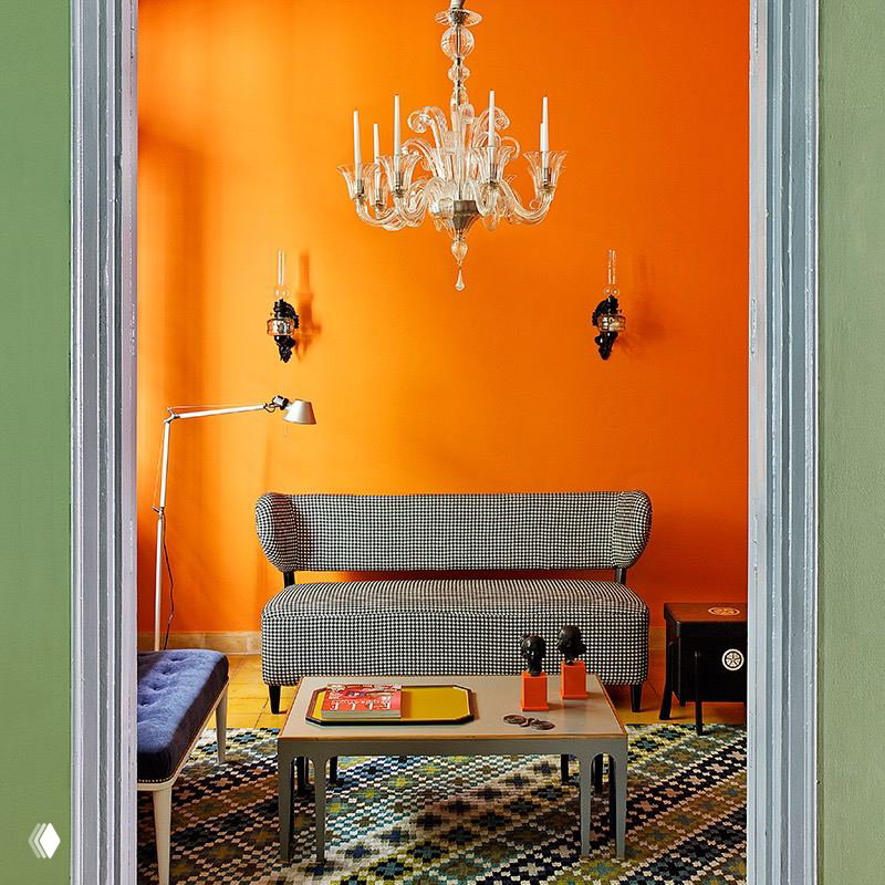 Ярко-оранжевая стена в гостиной с маленькой классической люстрой и винтажной мебелью — пример смелого цветового сочетания с традиционной люстрой.
