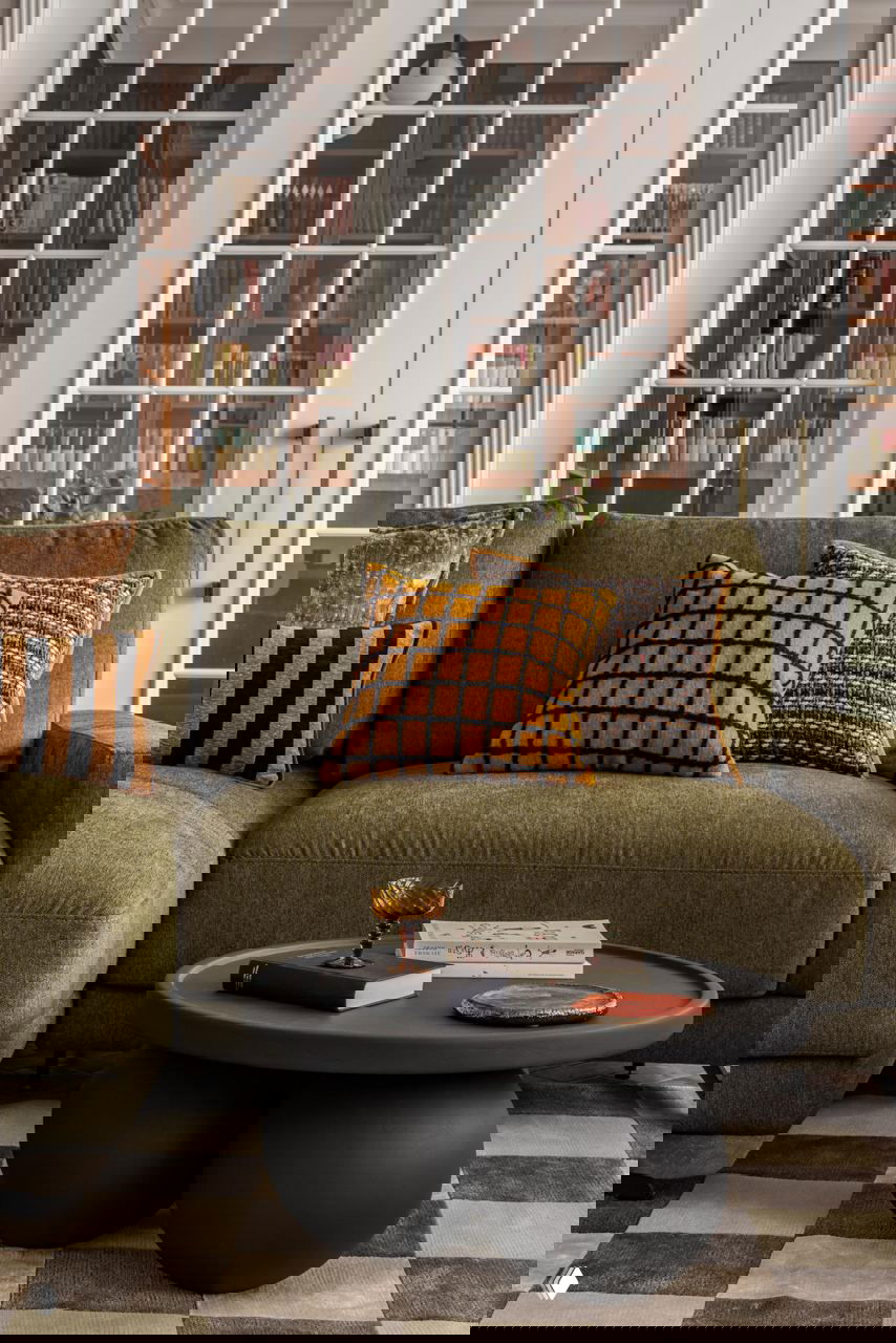 Гостиная с оливково-зелёным диваном, декоративными подушками в горчичных и коричневых тонах и низким круглым чёрным кофейным столиком на ковре.