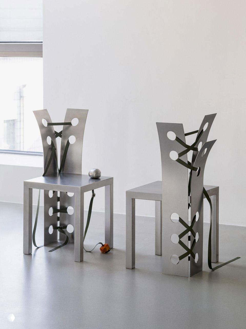 Два стула из нержавеющей стали вблизи у окна: изогнутые спинки с круглыми отверстиями и кожаными ремнями, которые визуально напоминают шнуровку.