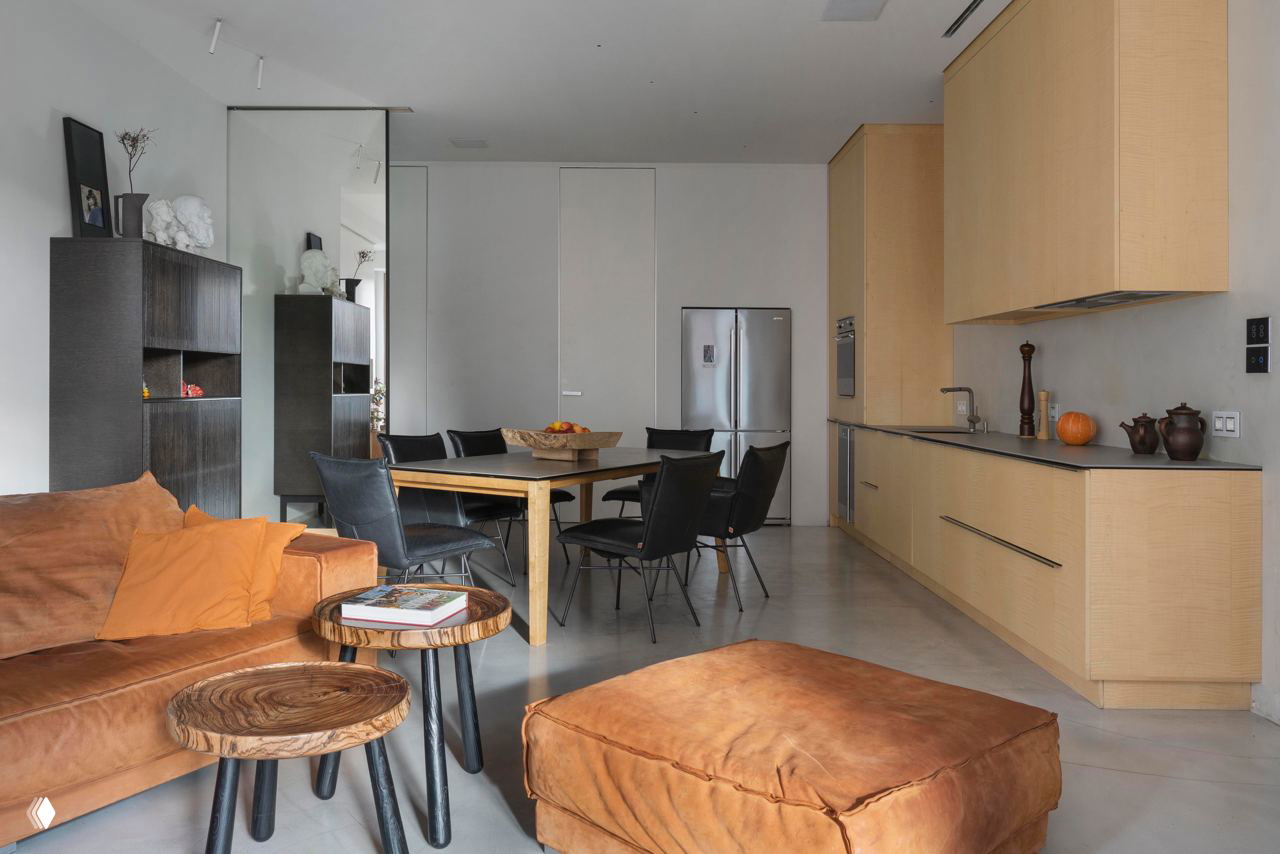 Кухонная и гостиная зона в едином пространстве: светлые кухонные фасады, деревянные поверхности и уютный диван коричневого тона в переднем плане.