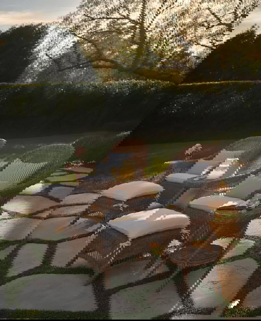 Набор садовой ротанговой мебели — кресла, стол и пуфы с белыми подушками на каменной террасе, снято при мягком вечернем свете.