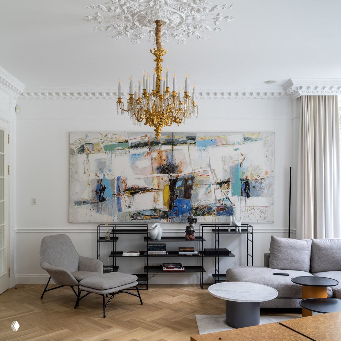 Светлый салон с белыми лепными деталями, золотая классическая люстра и большое абстрактное полотно на стене как акцентный элемент интерьера.