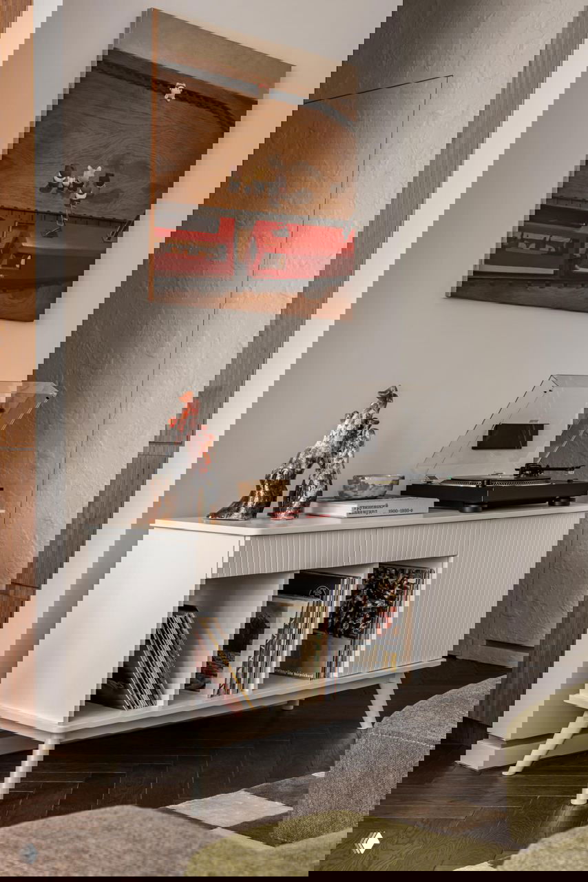 Низкий белый комод с проигрывателем и коллекцией виниловых пластинок, на стене картина и декоративные предметы, создающие ретро-атмосферу.