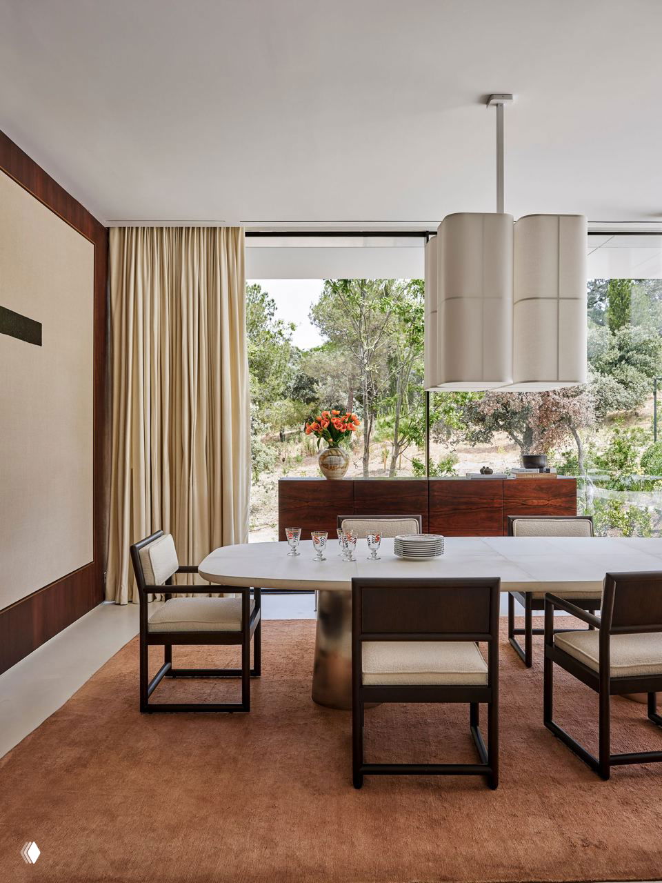 Столовая с овальным столом и мягкими креслами у панорамного окна, вид на сад, тёплые ткани и деревянные фактуры в пространстве.