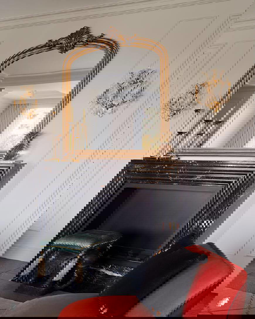 Чёрный мраморный камин с выраженной фактурой и позолоченным зеркалом над ним; интерьер в классическом стиле с декоративными настенными бра.